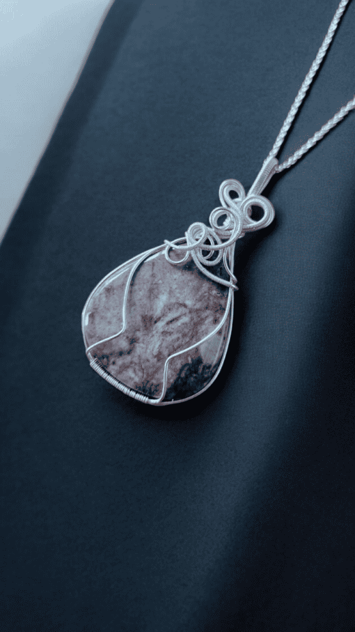 Close-up of a pendant necklace with an intricate silver wire design encasing a teardrop-shaped purple stone, on a dark fabric background.