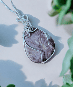 Silver wire-wrapped pendant featuring a marbled purple stone, surrounded by out-of-focus green plants in the foreground.