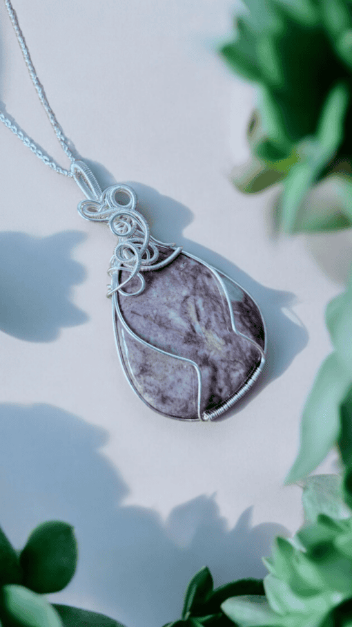 Silver wire-wrapped pendant featuring a marbled purple stone, surrounded by out-of-focus green plants in the foreground.