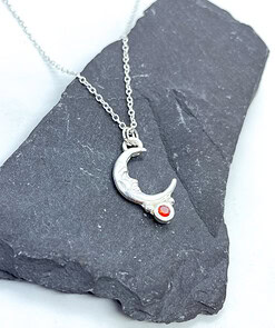 Silver crescent moon necklace with intricate details and a small red gemstone, displayed on a dark slate background.