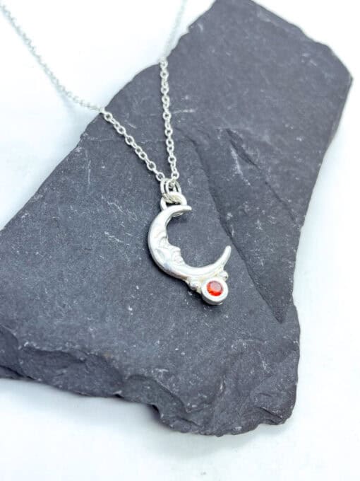 Silver crescent moon necklace with intricate details and a small red gemstone, displayed on a dark slate background.