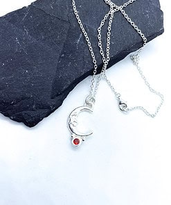 Silver crescent moon necklace with a red gemstone, displayed on a dark stone background with a white surface.