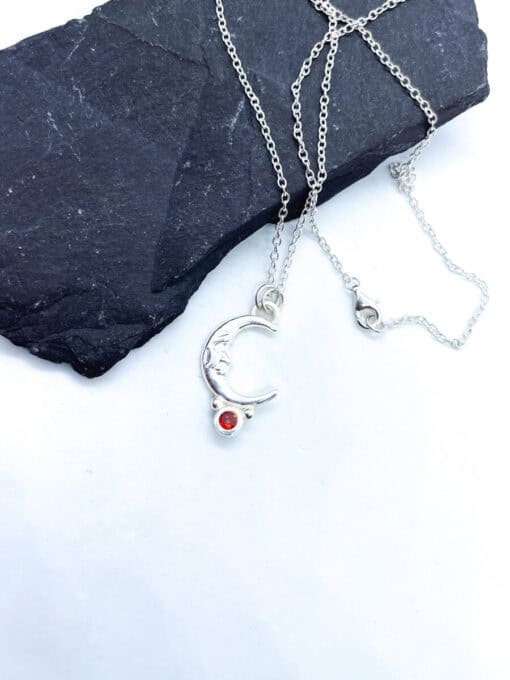 Silver crescent moon necklace with a red gemstone, displayed on a dark stone background with a white surface.