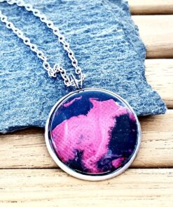 Round pendant with pink and black swirls on silver chain, placed on textured stone background.