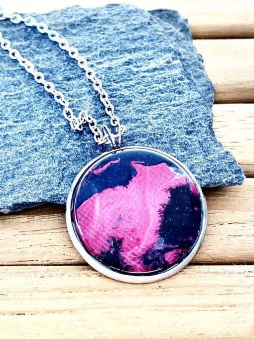 Round pendant with pink and black swirls on silver chain, placed on textured stone background.