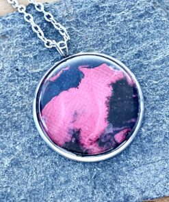 Round pendant with pink and black abstract design on stone background, attached to a silver chain.