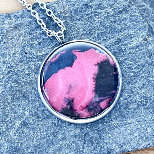 Round pendant with pink and black abstract design on stone background, attached to a silver chain.