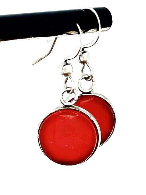 Two red circular earrings with silver edges and hooks, hanging on a black rod.