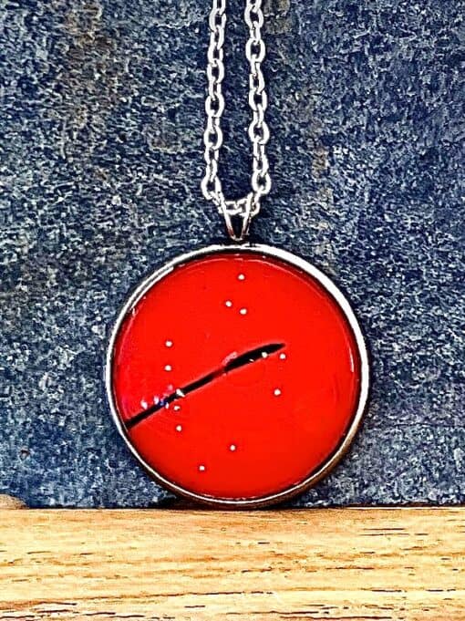 Red circular pendant with a black line and small white dots, hanging on a silver chain against a textured dark background.