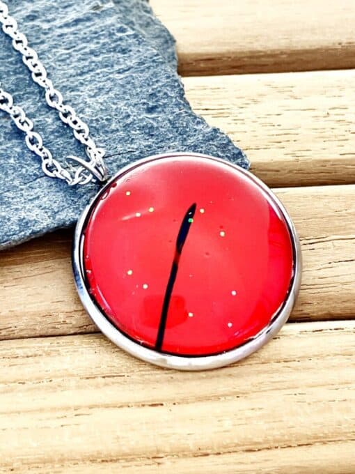 Red pendant with black line detail on silver chain, placed on gray stone, wooden background.