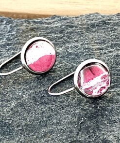 Silver round earrings with red and white marble-like design on a textured dark surface.