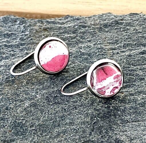 Silver round earrings with red and white marble-like design on a textured dark surface.