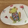 A hand-painted rock featuring a colorful frog, a dragonfly, a ladybug, and flowers on a wooden surface.