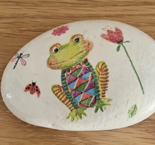 A hand-painted rock featuring a colorful frog, a dragonfly, a ladybug, and flowers on a wooden surface.