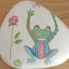 Painted rock with a colorful frog wearing striped clothes, a rose, and a dragonfly, placed on a wooden surface.