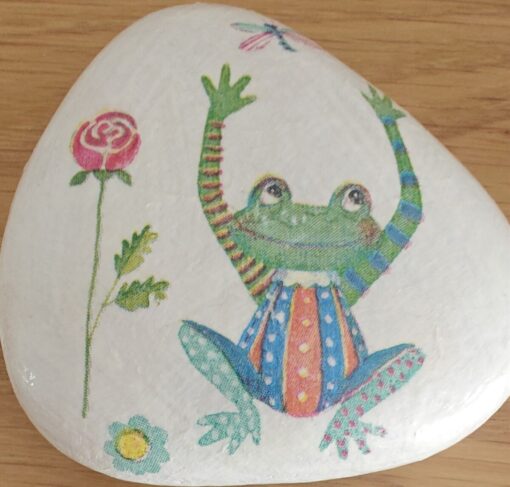 Painted rock with a colorful frog wearing striped clothes, a rose, and a dragonfly, placed on a wooden surface.