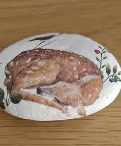 A painted rock featuring a detailed illustration of a sleeping fawn surrounded by foliage on a wooden surface.