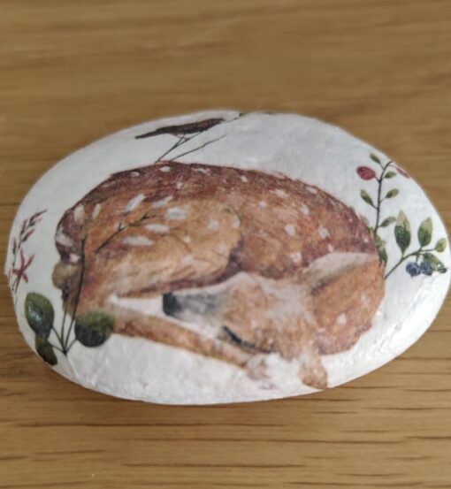 A painted rock featuring a detailed illustration of a sleeping fawn surrounded by foliage on a wooden surface.