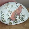 A painted rock featuring a fox, greenery, a bird, and a snail on a wooden surface.