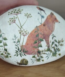 A painted rock featuring a fox, greenery, a bird, and a snail on a wooden surface.