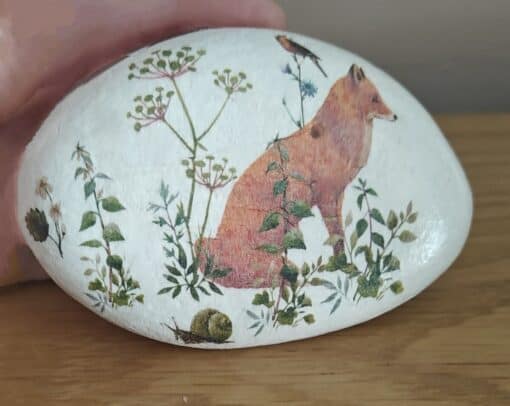 A painted rock featuring a fox, greenery, a bird, and a snail on a wooden surface.