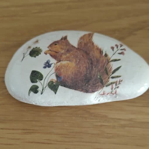 Smooth white stone with a painted image of a brown squirrel surrounded by green leaves and purple flowers.