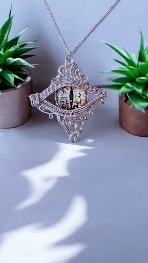 Silver wire-wrapped pendant with an eye design suspended between two small potted plants on a light surface.