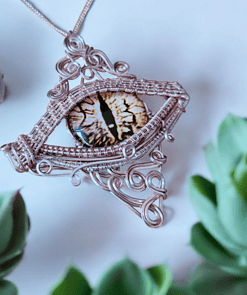 A close-up of a wire-wrapped pendant featuring a realistic eye design, surrounded by green succulent plants.