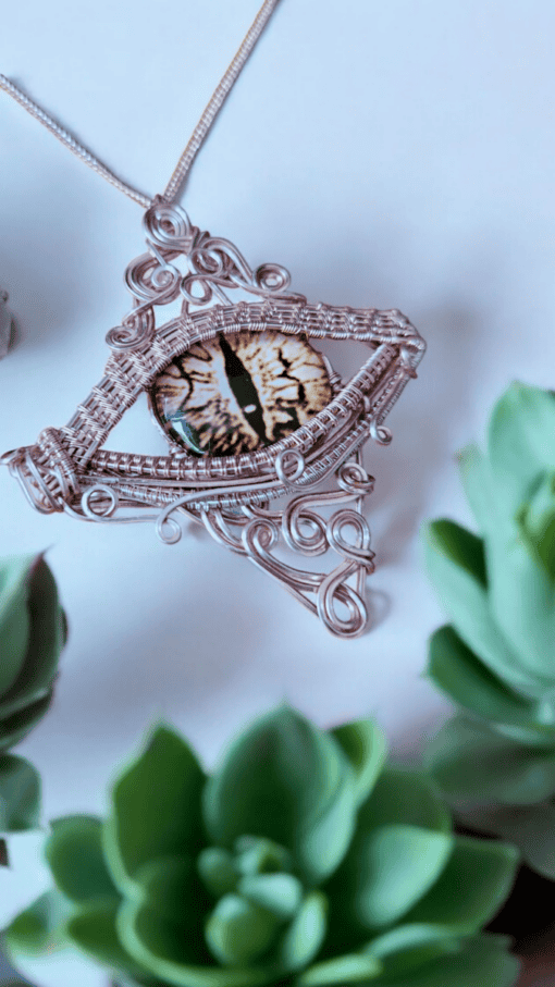 A close-up of a wire-wrapped pendant featuring a realistic eye design, surrounded by green succulent plants.