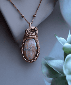 Handmade wire-wrapped stone jewellery pendant on a copper chain, displayed on a mannequin with green leaves in the background.