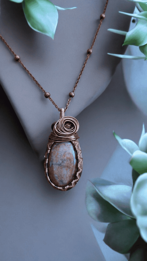 Handmade wire-wrapped stone jewellery pendant on a copper chain, displayed on a mannequin with green leaves in the background.