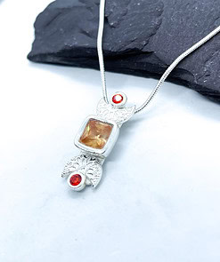 Silver necklace pendant with an orange square gemstone and two smaller round red gemstones, displayed on a light background.