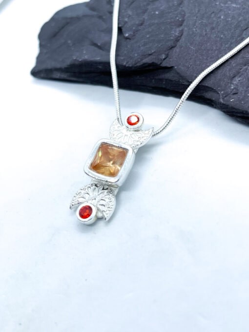 Silver necklace pendant with an orange square gemstone and two smaller round red gemstones, displayed on a light background.