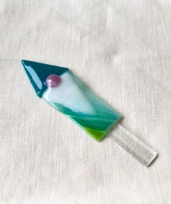 Glass rocket-shaped ornament with teal, white, and green colors, featuring a small pink dot, placed on a beige fabric background.