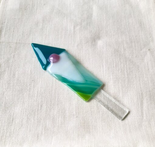 Glass rocket-shaped ornament with teal, white, and green colors, featuring a small pink dot, placed on a beige fabric background.