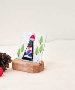 Glass art of a penguin with Santa hat on wooden stand, surrounded by pine cones and red ornament.