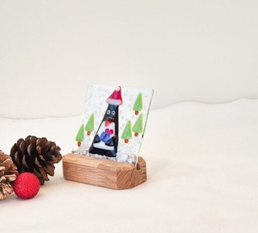 Glass art of a penguin with Santa hat on wooden stand, surrounded by pine cones and red ornament.