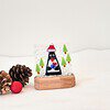 Glass penguin ornament with trees on wooden stand, surrounded by pinecones and a red glitter ball on white background.