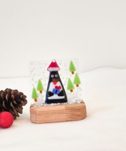 Glass penguin ornament with trees on wooden stand, surrounded by pinecones and a red glitter ball on white background.