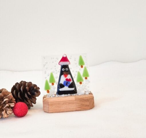 Glass penguin ornament with trees on wooden stand, surrounded by pinecones and a red glitter ball on white background.