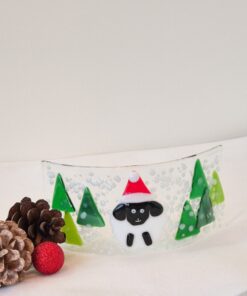 Glass artwork with a sheep in a Santa hat surrounded by green trees, pinecones, and a red ornament on a white surface.