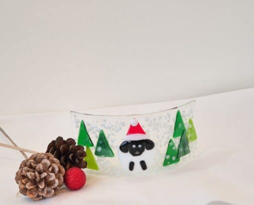 Glass artwork with a sheep in a Santa hat surrounded by green trees, pinecones, and a red ornament on a white surface.