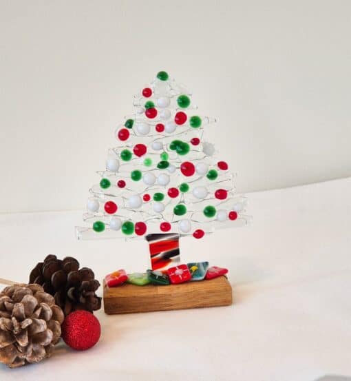 Glass Christmas tree with red, green, and white decorations on a wooden base, surrounded by pinecones and a red ornament.