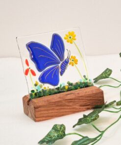 Stained glass with a blue butterfly and yellow flowers on a wooden stand, next to a vine on a white surface.