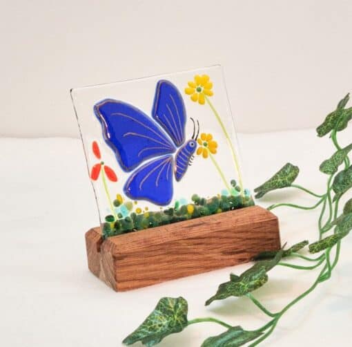 Stained glass with a blue butterfly and yellow flowers on a wooden stand, next to a vine on a white surface.