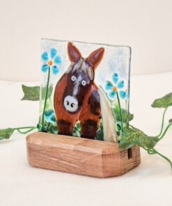 Glass art of a donkey with blue flowers on a wooden stand, surrounded by green leaves on a white background.