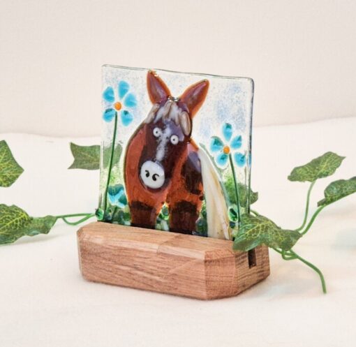 Glass art of a donkey with blue flowers on a wooden stand, surrounded by green leaves on a white background.