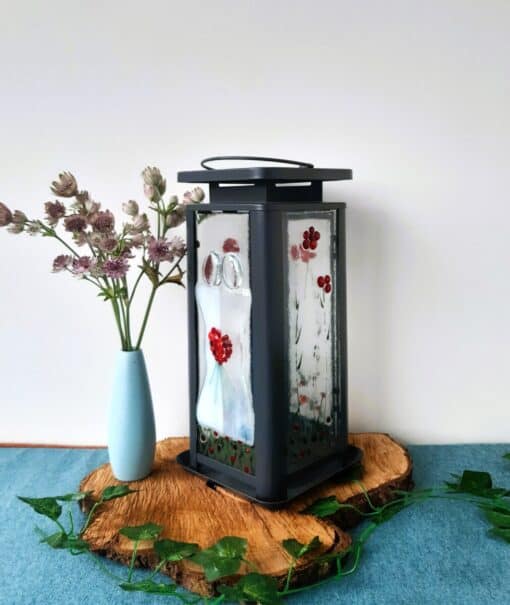 Black lantern with painted glass panel depicting a couple, next to a blue vase with flowers, on a wooden slice with green leaves.