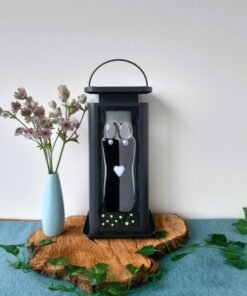 A blue vase with pink flowers sits next to a black lantern with artistic decorations on a wooden slab with green leaves.