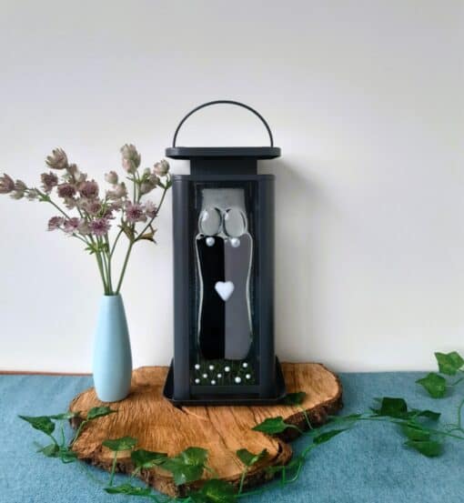 A blue vase with pink flowers sits next to a black lantern with artistic decorations on a wooden slab with green leaves.